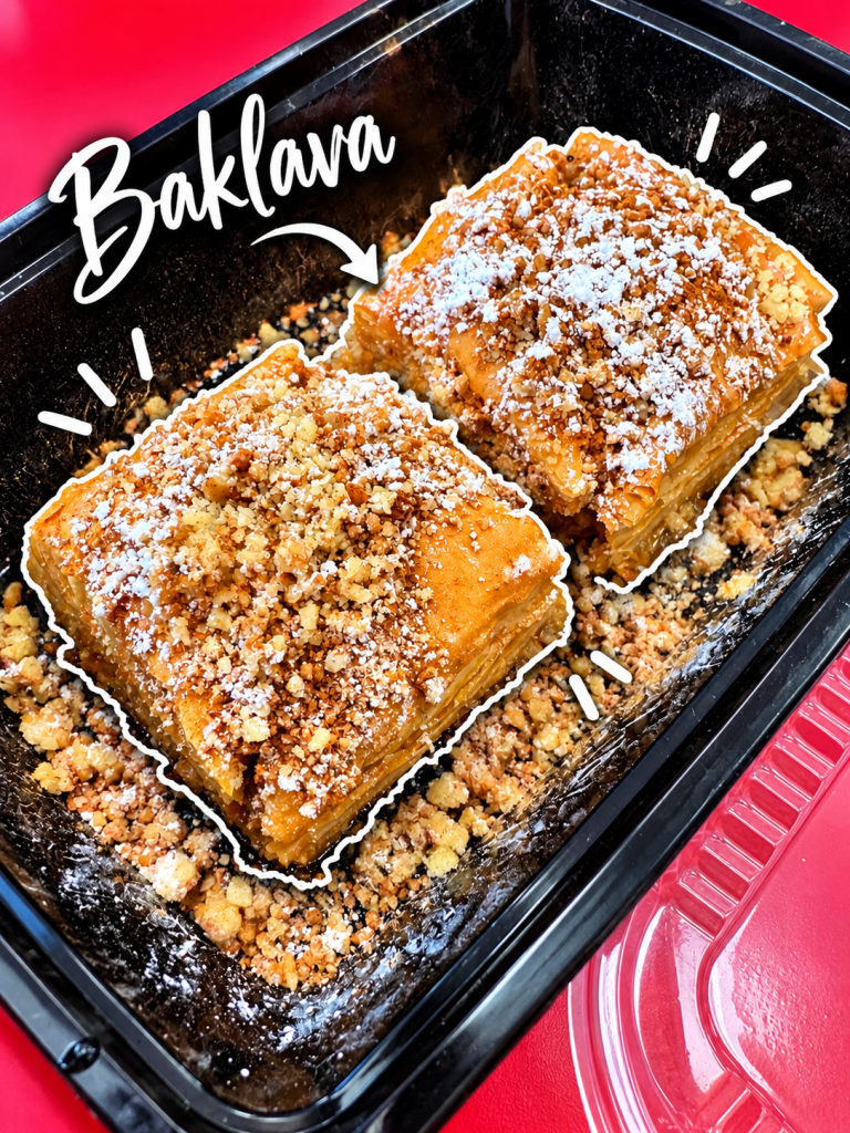 Close-up of two pieces of baklava topped with crushed nuts and powdered sugar in a black container.