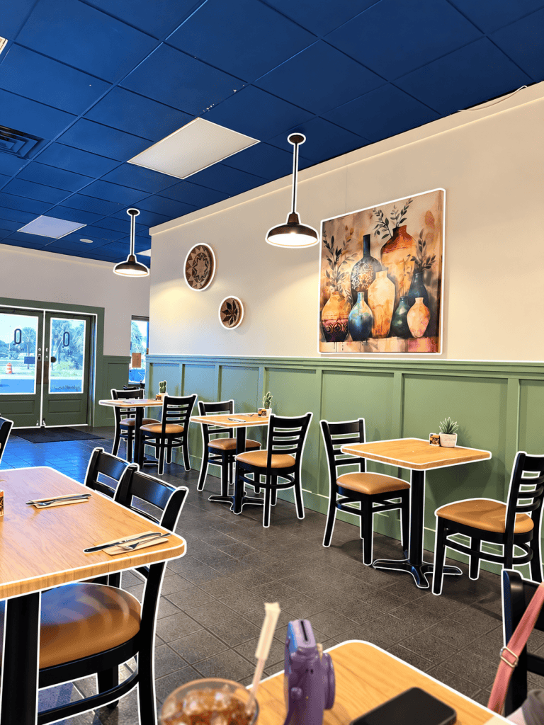 Interior of a restaurant featuring wooden tables and black chairs, decorated with a large painting of vases on the wall and pendant lighting hanging from a blue ceiling.