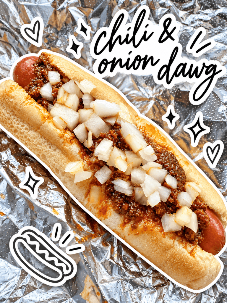 A chili and onion hot dog served in a soft bun, topped with chopped onions, on a silver foil background.