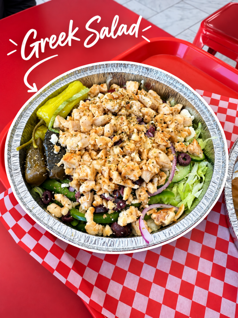 A close-up view of a Greek salad in a foil container, featuring chopped chicken, green peppers, olives, lettuce, and sprinkled with feta cheese. The salad is accompanied by a side of pickled peppers, with a vibrant red and white checkered background.