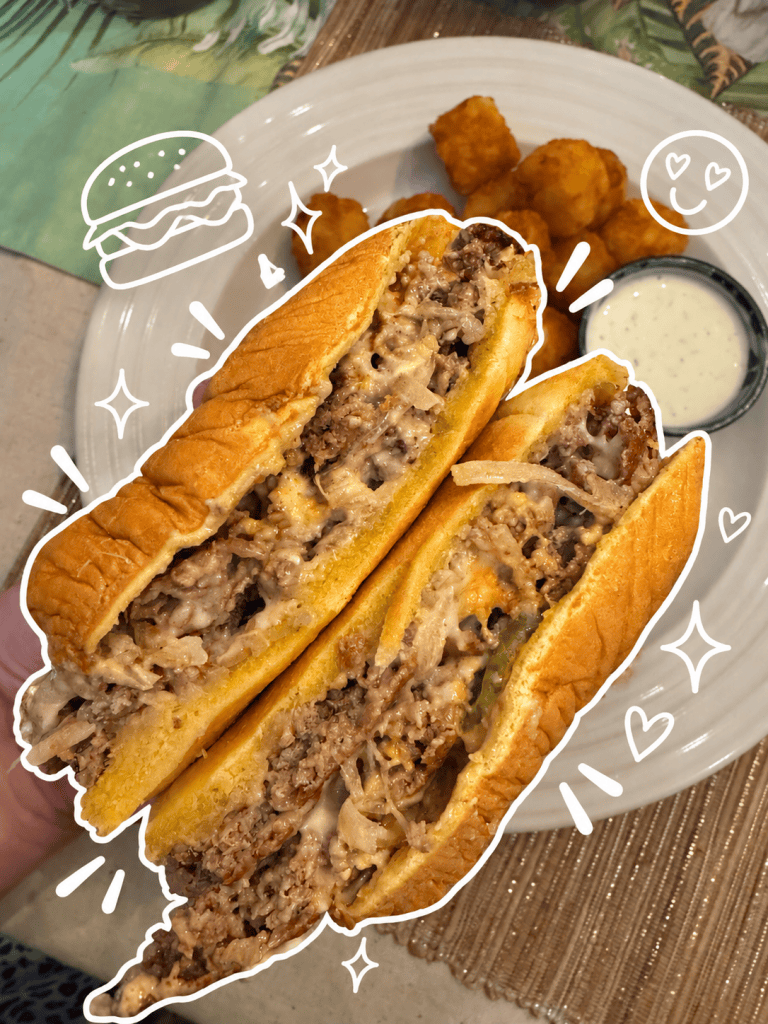 Close-up of a sliced cheesesteak sandwich with sautéed onions and sauce, served alongside crispy tater tots and a small cup of dipping sauce.