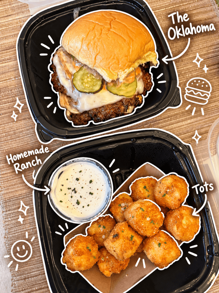 A close-up view of 'The Oklahoma' burger featuring a beef patty, melted cheese, pickles, and sauce, topped with a soft bun. Next to it, a portion of crispy tater tots and a small cup of homemade ranch dressing.