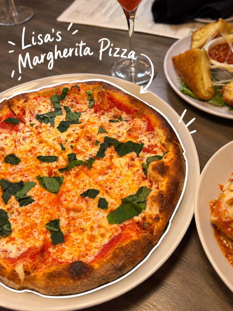 A Margherita pizza topped with fresh basil and melted cheese, served on a white plate. In the background, a cocktail glass and a plate of appetizers are visible.