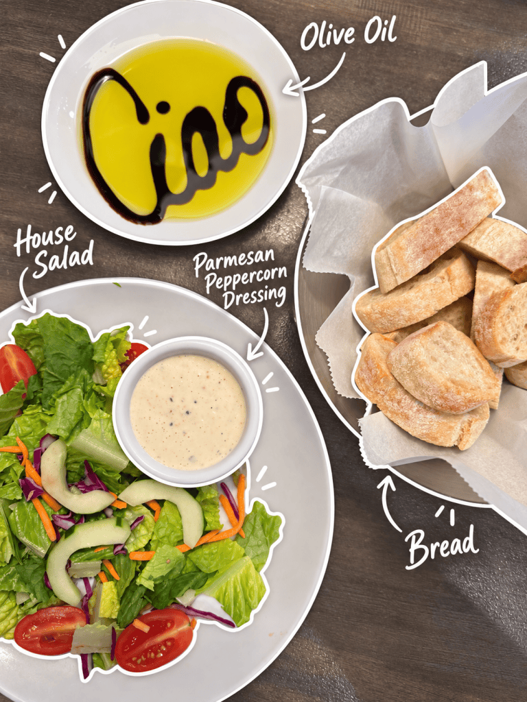 A bowl of olive oil with 'Ciao' written in it, a plate of house salad with various vegetables, a small bowl of Parmesan peppercorn dressing, and a basket of bread rolls.